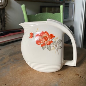 May include: A white ceramic pitcher with a floral design featuring orange and green flowers. The pitcher has a rounded body and a handle.