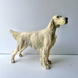 May include: A cream-colored, vintage-style dog figurine. The dog is standing with its head held high, featuring black spots on its legs and face. The sculpture has a textured surface and is likely made of ceramic or a similar material.
