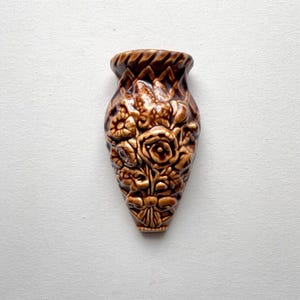 May include: A brown ceramic wall vase with a textured surface. The vase is shaped like a teardrop and features a relief design of roses and other flowers. The top rim has a textured, geometric pattern.