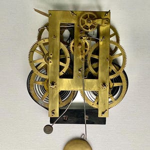 May include: A brass clock movement mechanism featuring a rectangular frame, multiple interlocking gears, and coiled springs. A thin metal rod extends downward, supporting a circular brass pendulum bob at the bottom, with a small weight attached.