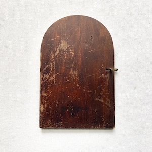 May include: A dark brown wooden door with an arched top, featuring a weathered surface with visible scratches and wear. A small, thin metal latch is attached to the right side of the door, extending slightly outward from the wooden frame.