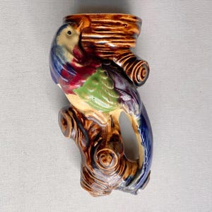 May include: A ceramic wall pocket in the shape of a colorful parrot perched on a brown tree branch. The bird features blue, red, green, and yellow hues. The branch has a textured, wood-grain appearance, and the pocket has an opening at the top.