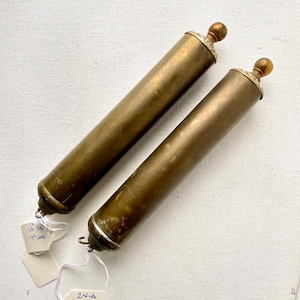 May include: Two antique brass cylinders with decorative finials. Each cylinder is approximately 9 inches long and 1.5 inches in diameter. The cylinders have a tarnished patina, and small tags are attached.