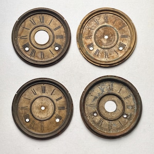 May include: Four antique clock faces with Roman numerals. The clock faces are circular, with a central hole and smaller holes for mounting. The metal has a weathered, aged appearance with a mix of brown and gold tones.