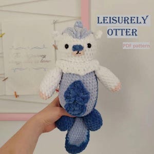 Puede incluir: Un juguete de peluche de una nutria tejido a crochet con un cuerpo blanco, una cola azul y detalles azules. El juguete está siendo sostenido en una mano. El texto "LEISURELY OTTER PDF pattern" es visible en la imagen.