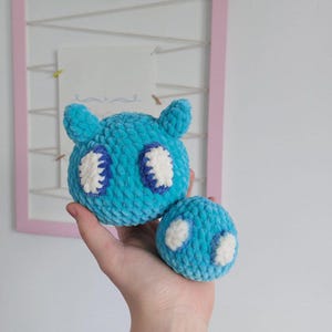 Puede incluir: Dos juguetes de peluche tejidos a crochet en forma de criatura con ojos grandes. El juguete más grande es azul con ojos blancos y el juguete más pequeño es azul con ojos blancos. Los juguetes se sostienen en una mano.