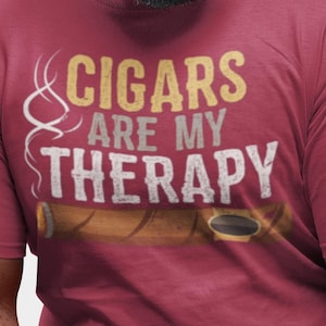 May include: Maroon t-shirt with the text "CIGARS ARE MY THERAPY" in gold and white. The design includes a cigar illustration. The shirt is worn by a person wearing blue jeans.