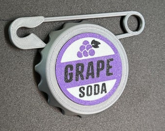 Puede incluir: Un pin gris impreso en 3D con un diseño de tapón de botella, que muestra las palabras "GRAPE SODA" y una ilustración de uva morada. El pin tiene un cierre de imperdible.