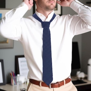 Può includere: Una cravatta blu navy a maglia è il punto focale di questa immagine. È abbinata a una camicia bianca e a una cintura in pelle marrone. La trama e il colore della cravatta contrastano con la camicia, creando un look classico e professionale. Il tema generale è l'abbigliamento da lavoro.