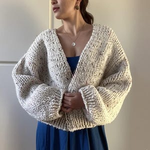 Puede incluir: Un cárdigan blanco con un patrón moteado y cuello en V. El cárdigan tiene mangas largas y un ajuste holgado.