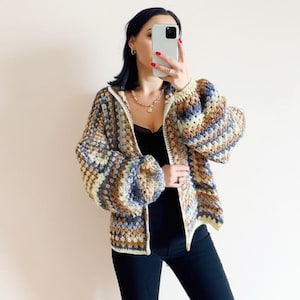 Może przedstawiać: Sweter dziany na drutach z zamkiem błyskawicznym i kolorowym paskowanym wzorem w odcieniach brązu, niebieskiego i kremowego. Sweter ma luźny krój i kaptur.