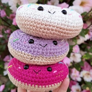 May include: A stack of three crocheted donut plushies. The top donut is white with rainbow sprinkles, the middle donut is purple with white sprinkles, and the bottom donut is pink with white sprinkles. Each donut has black eyes and a smiling mouth.