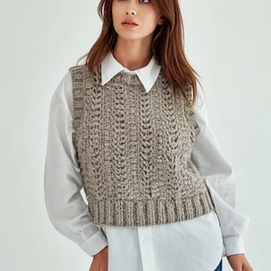 May include: A brown knitted waistcoat with a cable knit pattern. The waistcoat is sleeveless and has a round neckline. The waistcoat is worn over a white button-down shirt.