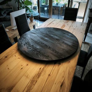 Könnte beinhalten: Ein großer, runder, dunkelgrauer Holz-Drehteller liegt auf einem hellen Esstisch aus Holz. Der Drehteller hat eine strukturierte Oberfläche und ist von schwarzen Stühlen umgeben. Der Tisch steht in einem hellen Raum mit Blick auf eine Außenterrasse.