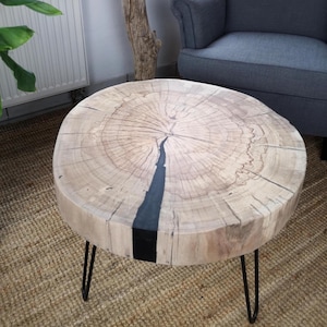 Op de afbeelding: Een ronde houten salontafel met een natuurlijke houtnerf afwerking. De tafel heeft een donker, zwart harsinlegsel en wordt ondersteund door drie zwarte metalen haarspeldpoten. De tafel heeft een diameter van ongeveer 46 cm.