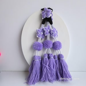 May include: A handcrafted, lavender-colored yarn garland with floral and tassel accents. The garland features pom-poms, beaded strands, and flower-shaped embellishments. The text "Heritage Loom" is visible on the bottom right.