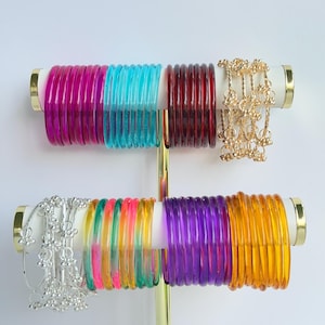 May include: A two-tiered gold jewelry stand displaying multiple sets of translucent bangles in magenta, turquoise, dark red, purple, and orange, alongside two sets of metallic bangles featuring small dangling charms in gold and silver tones.