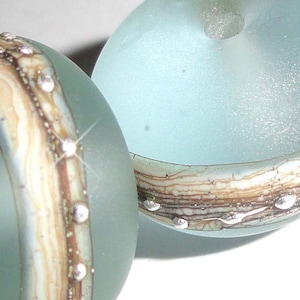 May include: Two glass beads with a light blue color and a brown and white swirl pattern. One bead has silver glitter embedded in the glass.