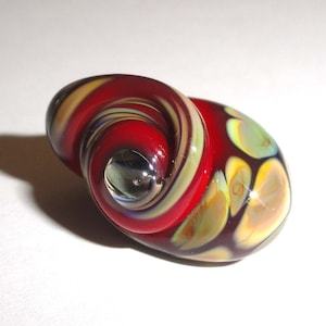 May include: A red, yellow, and green glass bead with a swirl pattern. The bead has a clear glass center.