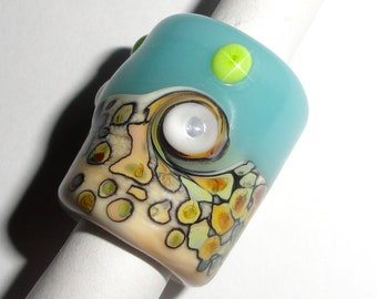 May include: A handmade glass ring with a unique design. The ring features a light blue top with a bright green dot, and a swirling pattern of white, yellow, and brown. The ring is mounted on a white cylindrical object.