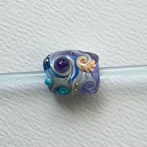 May include: A handcrafted glass bead with a sea-themed design. The bead features swirling blue, white, and purple colors, with a seashell and a purple gem. Small silver dots add detail. The bead is on a clear rod.