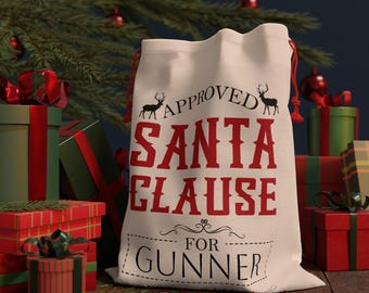 May include: A cream-colored Santa Claus gift bag with red text that reads "APPROVED SANTA CLAUSE FOR GUNNER." The bag has a drawstring closure and is surrounded by wrapped gifts and a Christmas tree.