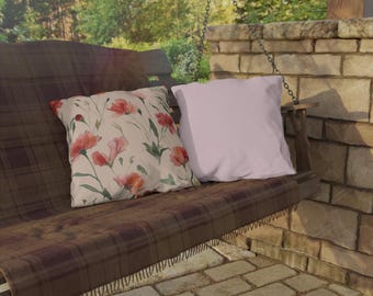 Pode incluir: Duas almofadas decorativas num baloiço de madeira. Uma almofada apresenta um padrão floral com flores vermelhas e rosa sobre um fundo creme. A outra almofada é de cor lilás claro lisa. O baloiço tem uma manta xadrez castanha.