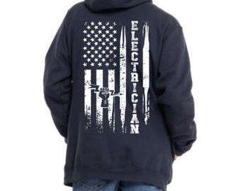 May include: Navy blue hoodie featuring a distressed American flag design with the word "ELECTRICIAN" in white. The graphic includes a lightning bolt and a fist. The hoodie has a hood and a front pocket.