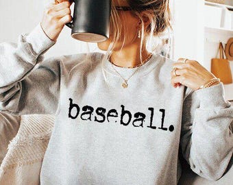 May include: A light gray sweatshirt with the word "baseball." printed in black, bold letters. The sweatshirt has long sleeves and a crew neck. The person is holding a black mug.