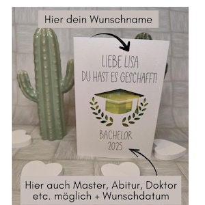 Peut inclure: Une carte de félicitations blanche avec le texte "LIEBE LISA DU HAST ES GESCHAFFT!" et "BACHELOR 2025". La carte présente un motif de chapeau de graduation et est ornée de feuilles de laurier. Un cactus vert et des décorations en forme de cœur sont en arrière-plan.