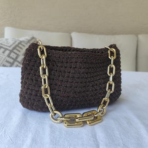 Könnte beinhalten: Eine braune Häkeltasche mit einem goldfarbenen Kettenträger. Die Tasche ist rechteckig mit einer strukturierten Oberfläche. Die Kette besteht aus großen, quadratischen Gliedern.