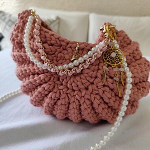 Könnte beinhalten: Staubrosa gehäkelte Handtasche mit Perlen- und Goldkettenträger. Die Tasche hat einen dekorativen Anhänger mit Traumfänger-Design, goldenen Federn und rosa Perlen. Die Tasche liegt auf einer weißen Oberfläche.