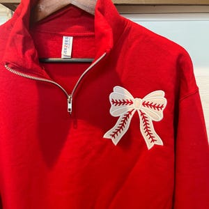 May include: A red quarter-zip sweatshirt with a white baseball bow applique. The sweatshirt has a zipper closure and a tag that reads "JERZEES". The bow is detailed with red stitching.