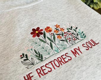 May include: Light gray sweatshirt with embroidered floral design in shades of red, orange, pink, and green. The phrase "HE RESTORES MY SOUL" is embroidered in red below the flowers. The sweatshirt is a casual, comfortable style.