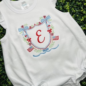 May include: White baby romper with a patriotic embroidered design. The design includes a red letter "E", blue bows, red and blue accents, and small American flags. The romper has ruffled sleeves and the text "CORB Blanks 3-6M".
