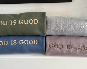 May include: Four folded sweatshirts in olive green, gray, blue, and mauve. Each sweatshirt has the embroidered text "GOD IS GOOD" in a contrasting color. The sweatshirts are stacked on a white shelf.