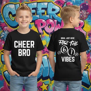 Peut inclure: T-shirts noirs avec texte blanc. L'un dit "CHEER BRO" devant. L'autre dit "BRUH, JUST HERE FOR THE 6 7 8 VIBES" au dos. Le fond est un mur de graffiti coloré.