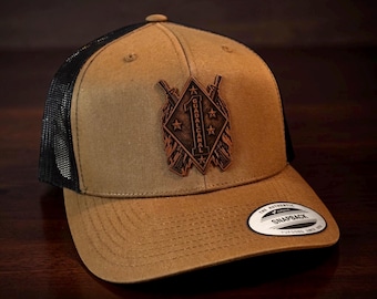 May include: A tan snapback cap with a black mesh back and a brown leather patch on the front. The patch features a diamond shape with the text GUADALCANAL, a number one, and stars, flanked by two crossed rifles. A round sticker is on the brim.