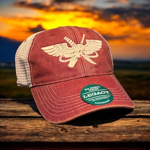 May include: Red and white trucker hat with a white skull and crossbones with wings design. The hat has a green patch that says "Old Favorite Trucker Legacy".