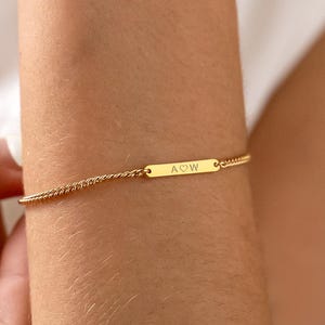 Può includere: Un braccialetto dorato con una catena delicata e una barretta rettangolare. La barretta è incisa con le lettere "AOW". Il braccialetto è indossato su un polso e la catena è finemente dettagliata.