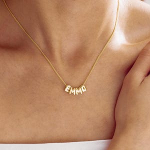 Puede incluir: Un collar de cadena en tono dorado con cuatro dijes metálicos individuales, redondeados, que forman el nombre EMMA. Las letras están suspendidas de una delicada cadena de eslabones finos que descansa sobre la clavícula.