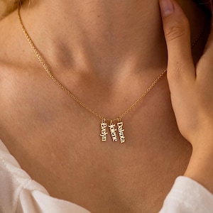 May include: A gold-coloured necklace featuring three vertical rectangular pendants hanging from a delicate chain. Each pendant is engraved with a name in a clear serif font, specifically the names Evelyn, Jolene, and Dakota, arranged side by side.
