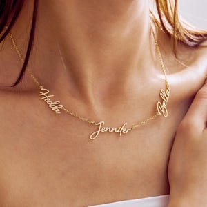 Peut inclure: Un collier doré avec trois prénoms : "Hedda", "Jennifer" et "Bella" en écriture cursive. Le collier est porté autour du cou, les noms étant affichés sur la poitrine. La chaîne est fine et les noms sont reliés.