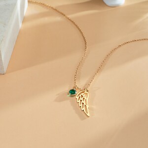 K&ouml;nnte beinhalten: Eine goldfarbene Halskette mit zarter Kette. Die Halskette hat einen kleinen, runden, gr&uuml;nen Edelstein und einen fl&uuml;gelf&ouml;rmigen Anh&auml;nger. Der Fl&uuml;gelanh&auml;nger hat ein offenes Design. Die Halskette wird auf einer hellbeigen Oberfl&auml;che pr&auml;sentiert.