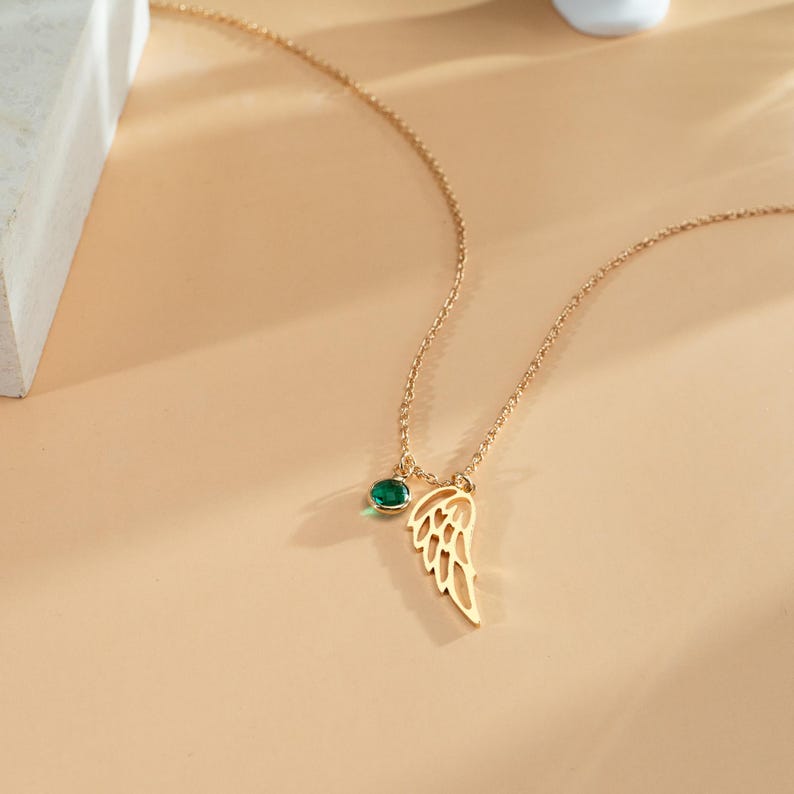 K&ouml;nnte beinhalten: Eine goldfarbene Halskette mit zarter Kette. Die Halskette hat einen kleinen, runden, gr&uuml;nen Edelstein und einen fl&uuml;gelf&ouml;rmigen Anh&auml;nger. Der Fl&uuml;gelanh&auml;nger hat ein offenes Design. Die Halskette wird auf einer hellbeigen Oberfl&auml;che pr&auml;sentiert.