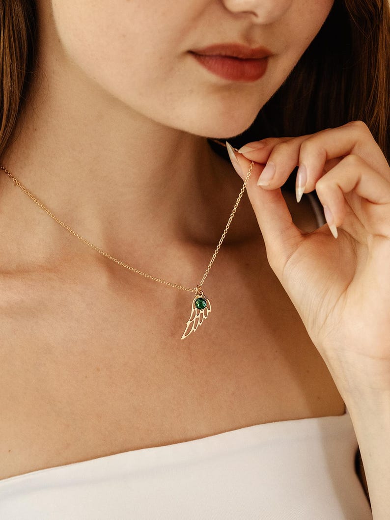 K&ouml;nnte beinhalten: Eine goldfarbene Halskette mit einem Fl&uuml;gelanh&auml;nger. Der Anh&auml;nger hat ein offenes Design und einen kleinen, runden, gr&uuml;nen Edelstein. Die Halskette wird pr&auml;sentiert, wobei die Kette und die Details des Anh&auml;ngers hervorgehoben werden.