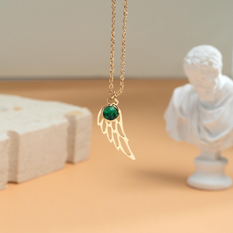K&ouml;nnte beinhalten: Eine goldfarbene Halskette mit einem fl&uuml;gelf&ouml;rmigen Anh&auml;nger mit einem gr&uuml;nen Edelstein. Die Kette h&auml;ngt vor einem neutralen Hintergrund, mit einer wei&szlig;en B&uuml;stenskulptur im Hintergrund. Der Anh&auml;nger ist etwa 2 cm lang.
