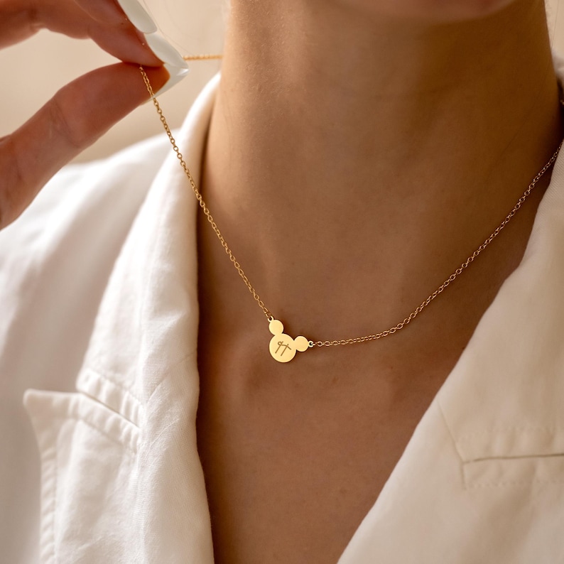 May include: A gold-coloured necklace featuring a pendant shaped like a cartoon character's head. The pendant has two small circles for ears and a larger circle with the initials "TT" engraved. The necklace is on a delicate chain.