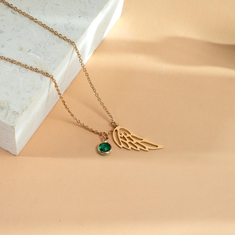 K&ouml;nnte beinhalten: Eine goldfarbene Halskette mit einem filigranen Fl&uuml;gelanh&auml;nger und einem kleinen, runden, smaragdgr&uuml;nen Edelstein. Die Kette hat eine feine Gliederkette und wird vor neutralem Hintergrund pr&auml;sentiert, um das Design und die Farbe hervorzuheben.