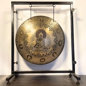 K&ouml;nnte beinhalten: Ein gro&szlig;er, runder, goldfarbener Gong mit aufwendigen Designs und einer Buddha-Figur in der Mitte, der an einem schwarzen Metallst&auml;nder h&auml;ngt.
